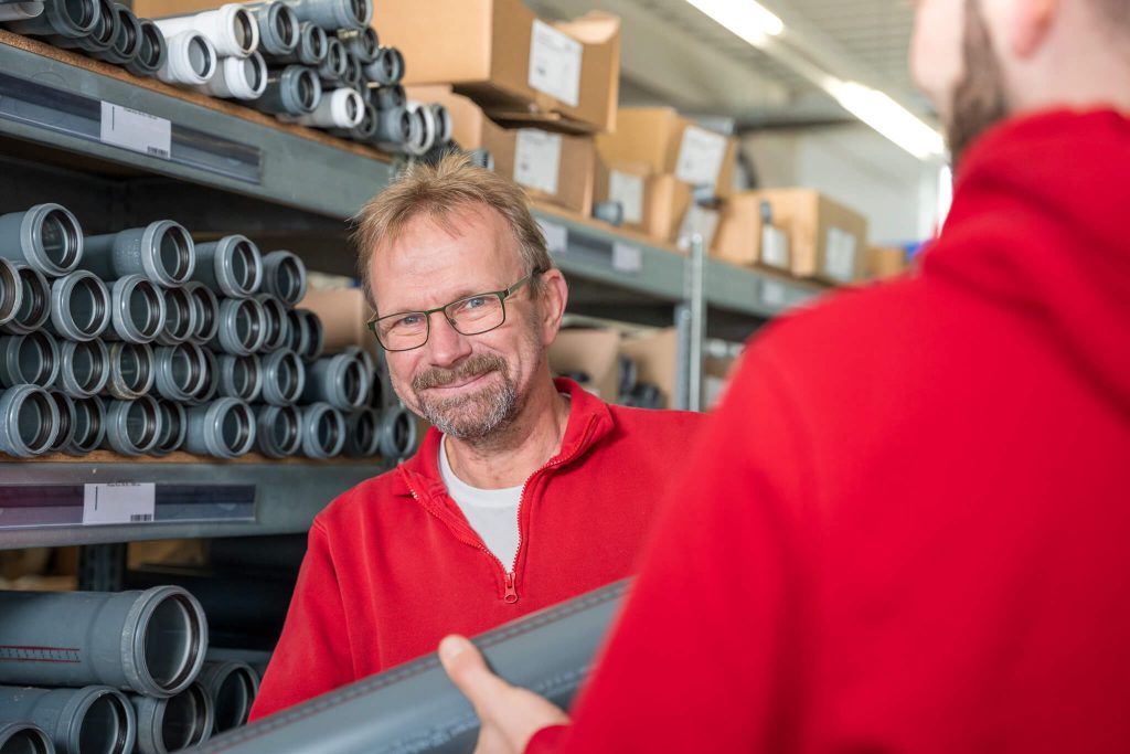 Freundlicher Mitarbeiter mit Brille vor Lagerregal mit grauen Abwasserrohren